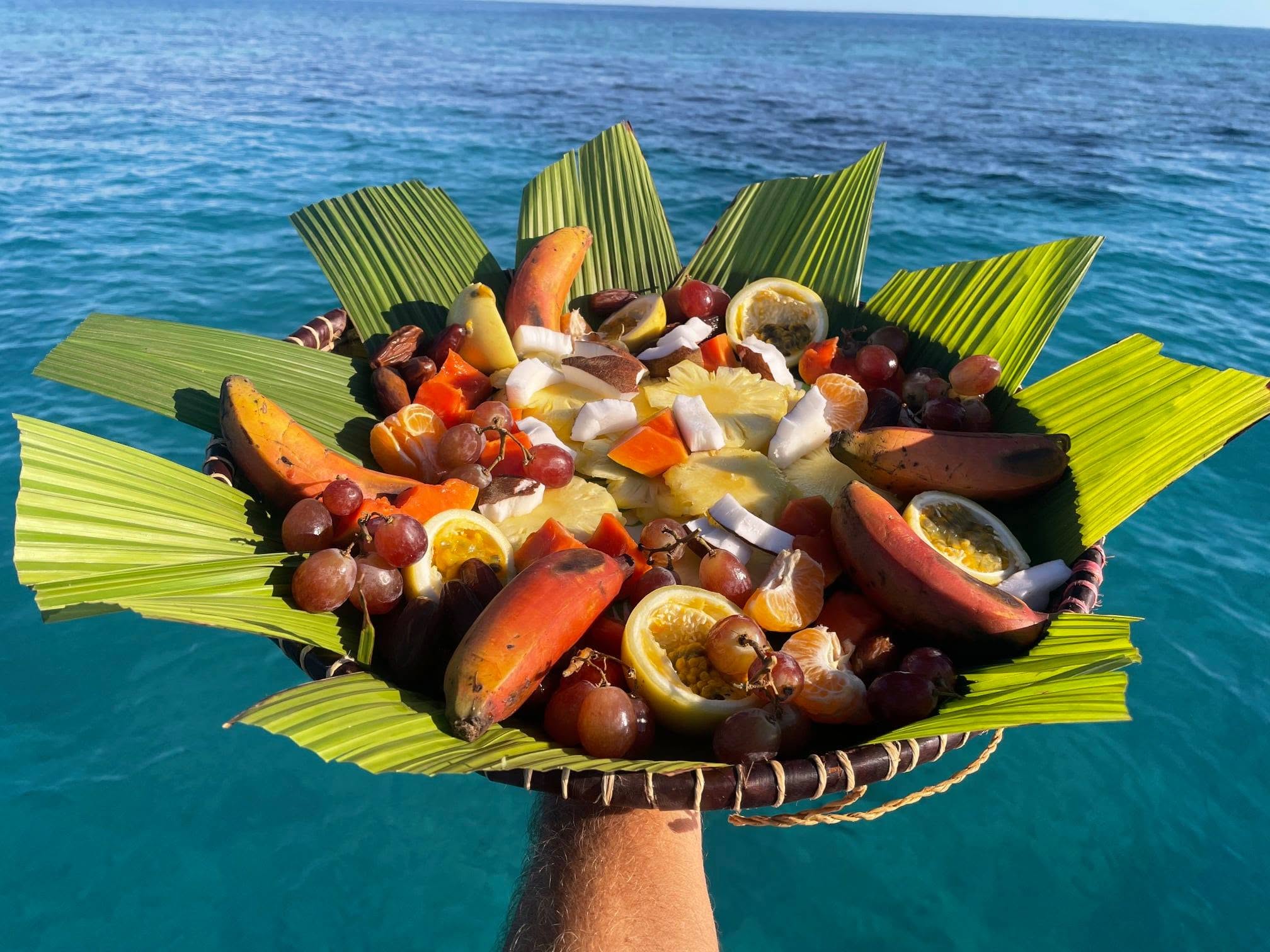 Plateau de fruits frais tressé de feuilles de palmier, présenté à bord face au lagon